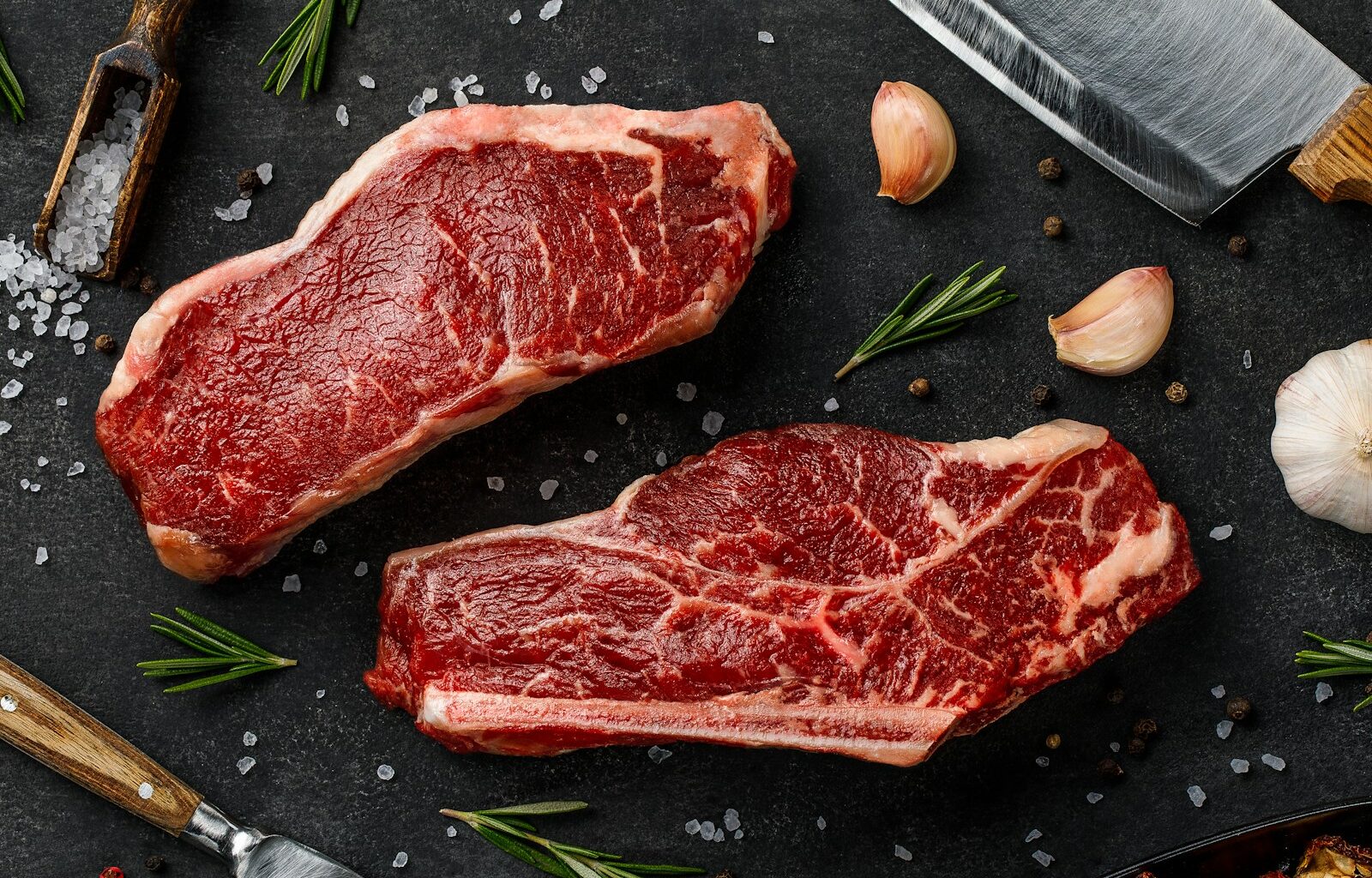 two steaks on a cutting board next to garlic, garlic, and a knife