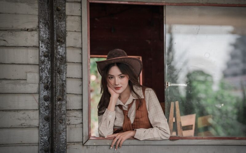 Pozycja kowbojka – jak ją wykonać, warianty i bezpieczeństwo 13 A woman in a vintage western outfit poses thoughtfully by a rustic window.