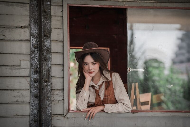 Pozycja kowbojka – jak ją wykonać, warianty i bezpieczeństwo 5 A woman in a vintage western outfit poses thoughtfully by a rustic window.